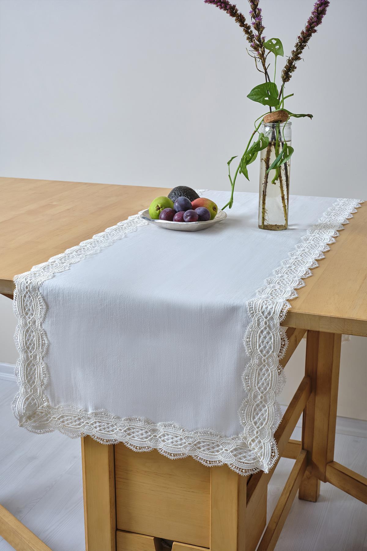 Camino de mesa blanco con detalles de encaje, con un frutero y un jarrón de cristal sobre una mesa de madera