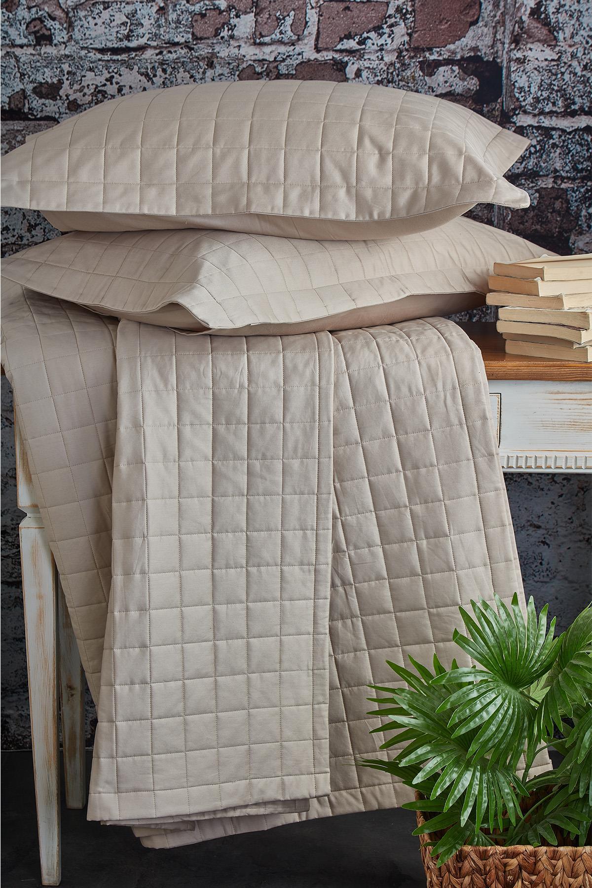 Colcha acolchada matrimonial, color crema, con dos almohadas y sobre una mesa