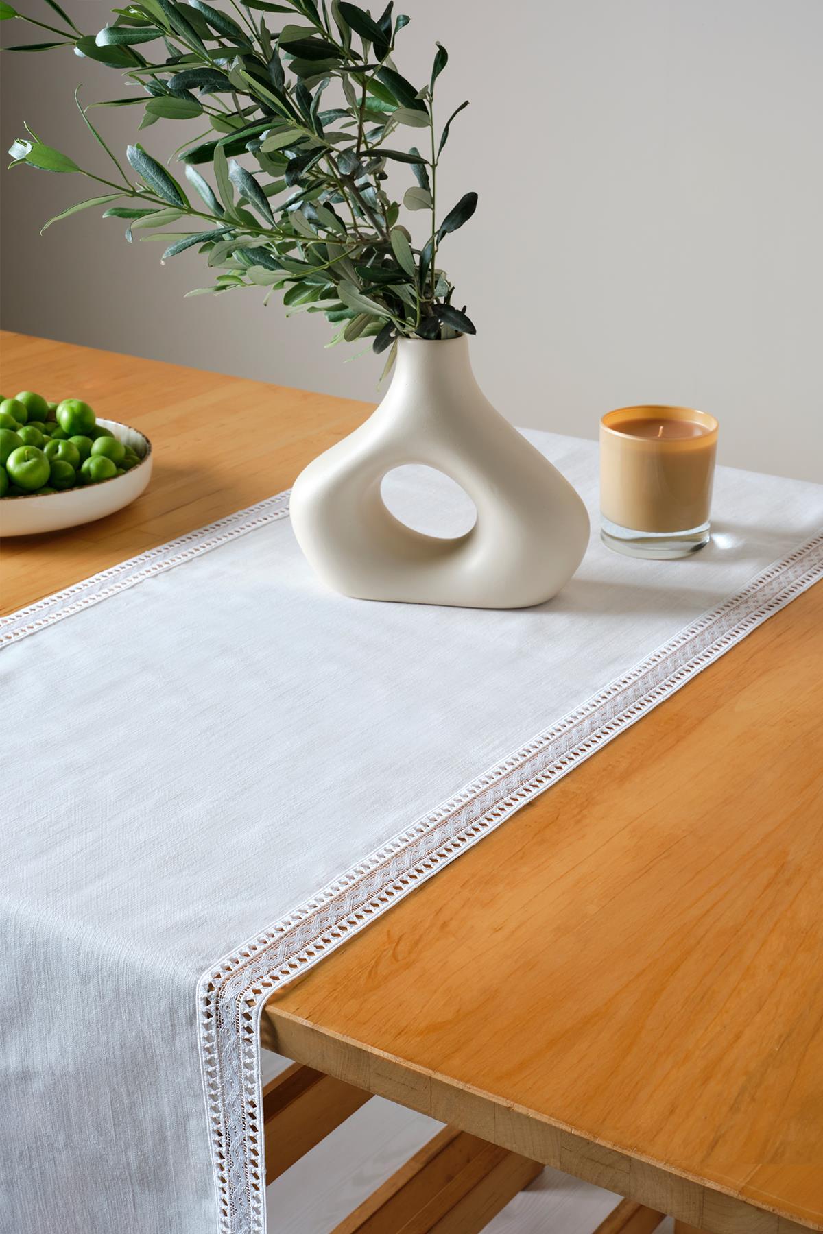 Camino de mesa de encaje blanco sobre una mesa de madera rectangular, con un jarrón moderno, frutas verdes y decoración con velas.