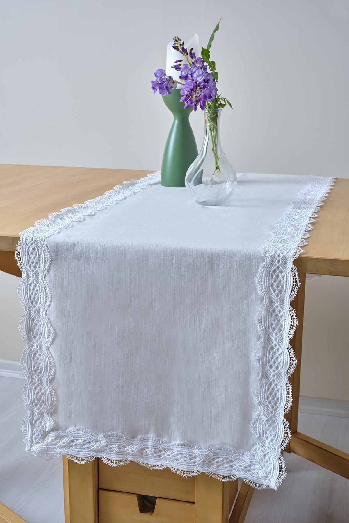 Camino de mesa blanco con borde de encaje, decorado con un jarrón de cristal y flores moradas sobre una mesa de madera