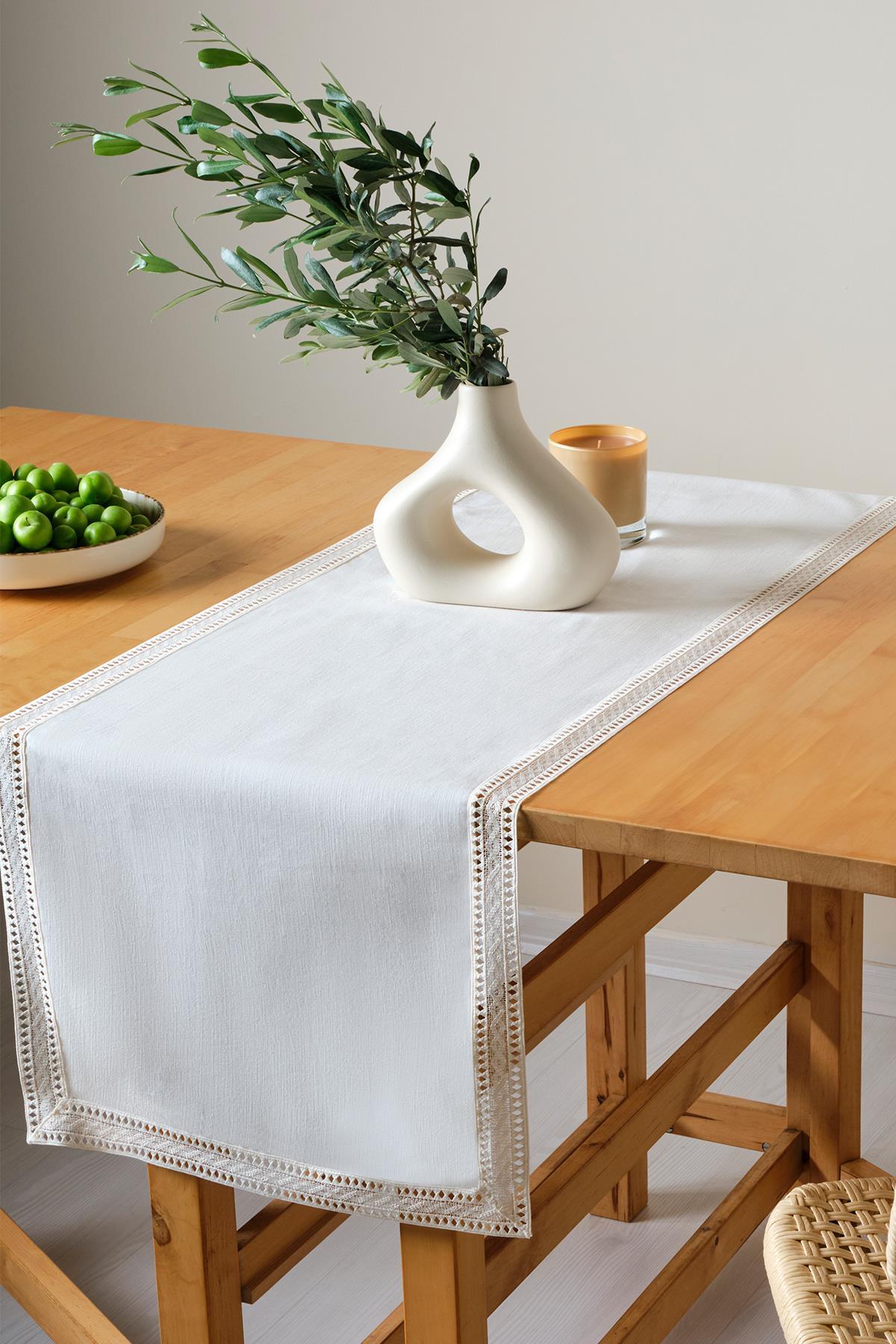 Mesa de madera decorada con un camino de mesa de encaje, con un jarrón moderno y ciruelas verdes en un plato