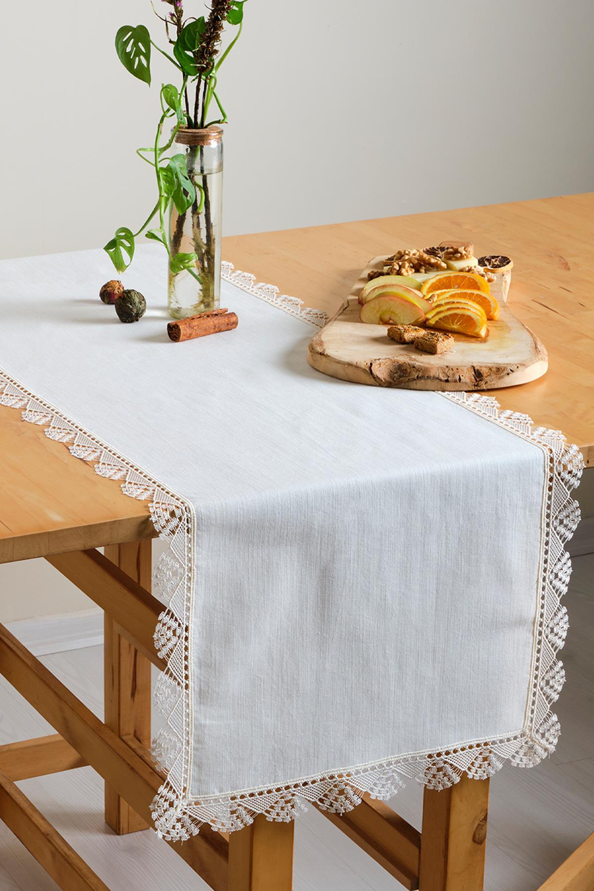 Camino de mesa blanco de encaje, mantel decorado con frutas y planta ornamental sobre una mesa de madera